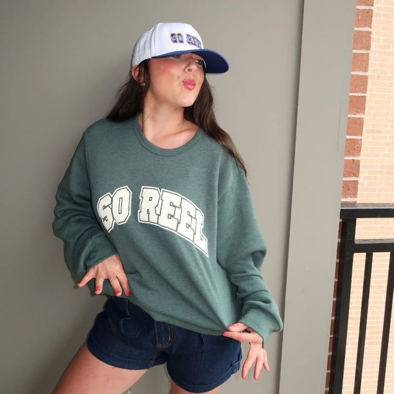 Load image into Gallery viewer, Person wearing a green 'SO REEL' sweatshirt and blue cap, standing against a neutral wall.